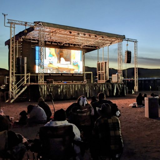 Stage with digital screen and speakers in a desert at night with a movie playing and people watching