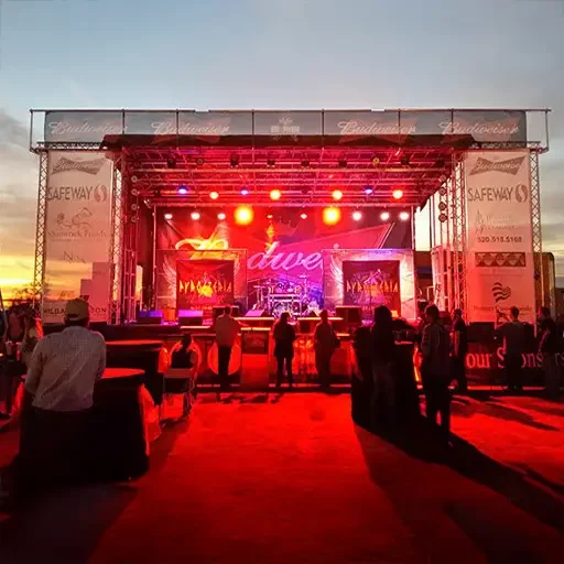Mobile 33′ x 20′ XXL stage, concert par cans and spot lighting, and Mitus speakers. 33' x 20' XXL stage with mesh graphics and warm concert lighting. People watching concert at sunset.